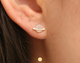 Puede incluir: Un pendiente de tono dorado con una pequeña piedra redonda brillante engastada en pavé en forma de planeta con una fina banda dorada alrededor del centro.