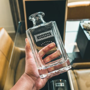 May include: A clear glass decanter with a square base and a label that reads "JACKSON RESERVE 1891".