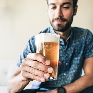 Puede incluir: Una persona sosteniendo un vaso de cerveza con la palabra "Sayers" grabada en el lateral. La cerveza es de color claro y tiene una cabeza blanca.