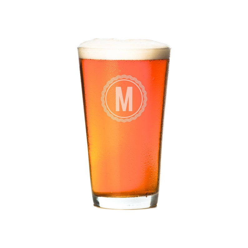 Puede incluir: Un vaso de pinta de vidrio transparente con un borde esmerilado lleno de una cerveza de color &aacute;mbar claro. El vaso tiene un c&iacute;rculo blanco con la letra "M" en el centro.