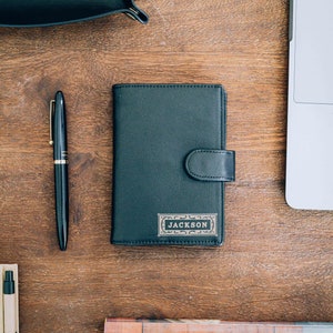 May include: A black leather wallet with a gold embossed name tag that reads "JACKSON". The wallet is on a wooden surface with a pen and a tray with the word "SMITH" embossed on it.