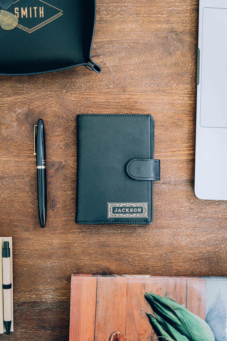 May include: A black leather wallet with a gold embossed name tag that reads "JACKSON". The wallet is on a wooden surface with a pen and a tray with the word "SMITH" embossed on it.