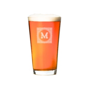 Puede incluir: Un vaso alto y transparente lleno de cerveza de color &aacute;mbar claro. El vaso tiene un dise&ntilde;o esmerilado con la letra "M" en un marco decorativo.