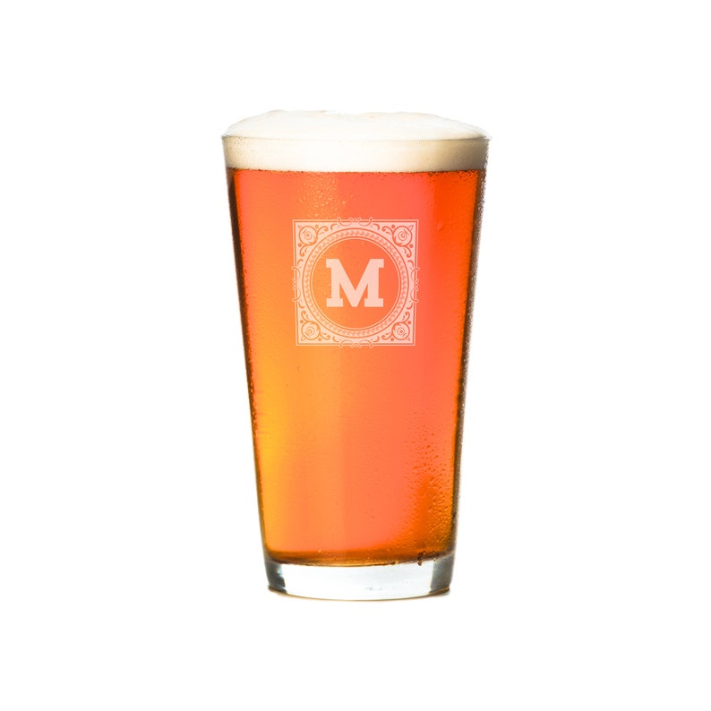 Puede incluir: Un vaso alto y transparente lleno de cerveza de color &aacute;mbar claro. El vaso tiene un dise&ntilde;o esmerilado con la letra "M" en un marco decorativo.