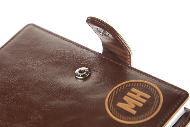 May include: A brown leather journal with a flap closure and a metal button. The journal has a personalised engraved wooden circle with the initials "MH" on the front.
