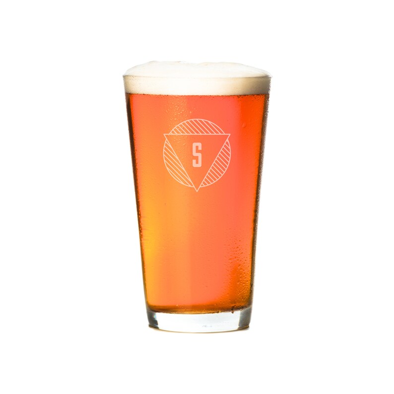 Puede incluir: Un vaso alto de cerveza de color &aacute;mbar con una cabeza blanca. El vaso tiene un logotipo con un c&iacute;rculo y un tri&aacute;ngulo en su interior, con la letra "S" en el centro.