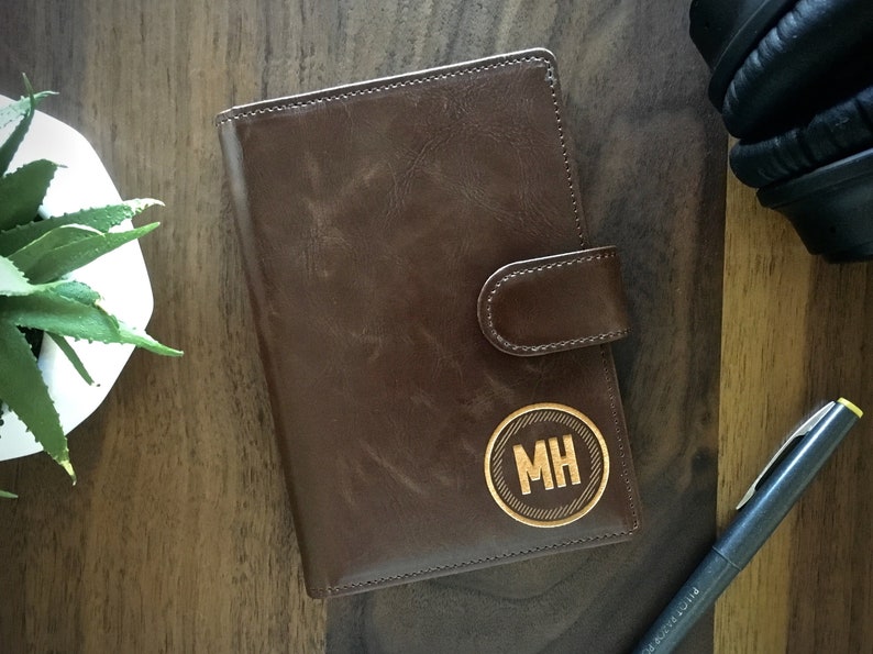 May include: A brown leather journal with a flap closure and a gold circle with the initials "MH" engraved on the front.