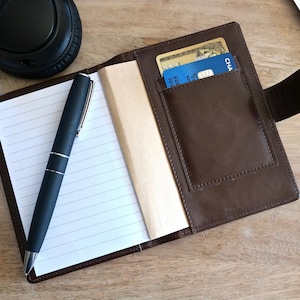 May include: A brown leather notebook with a pen and a credit card holder. The notebook has lined pages and a brown leather cover. The credit card holder is on the right side of the notebook and has a flap that closes with a snap.