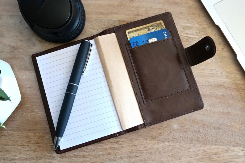 May include: A brown leather notebook with a pen and a credit card holder. The notebook has lined pages and a brown leather cover. The credit card holder is on the right side of the notebook and has a flap that closes with a snap.