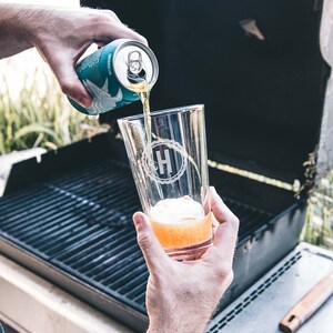 Puede incluir: Una persona vierte una lata de cerveza azul claro en un vaso transparente con un monograma blanco "H" en &eacute;l. El vaso est&aacute; sobre una parrilla negra.