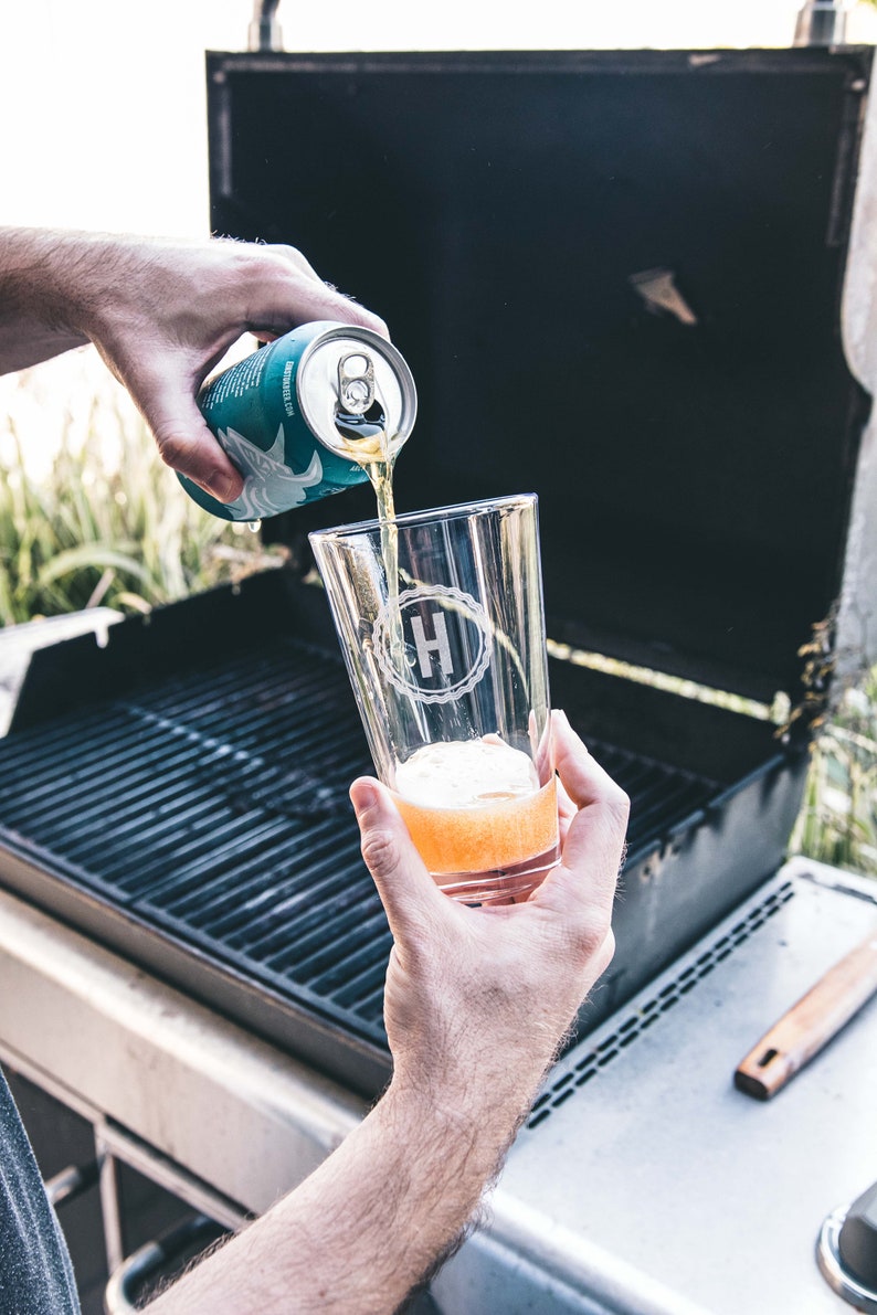 Puede incluir: Una persona vierte una lata de cerveza azul claro en un vaso transparente con un monograma blanco "H" en &eacute;l. El vaso est&aacute; sobre una parrilla negra.
