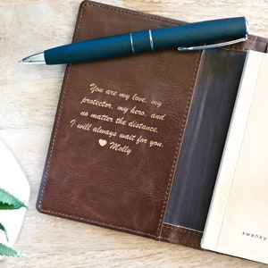 May include: A brown leather journal cover with a gold engraved message: "You are my love, my protector, my hero, and no matter the distance. I will always wait for you. Molly". The cover has a pocket for a pen and a black pen is placed in the pocket. The journal is open to a blank page with the words "swanky badger" printed on the bottom right corner.