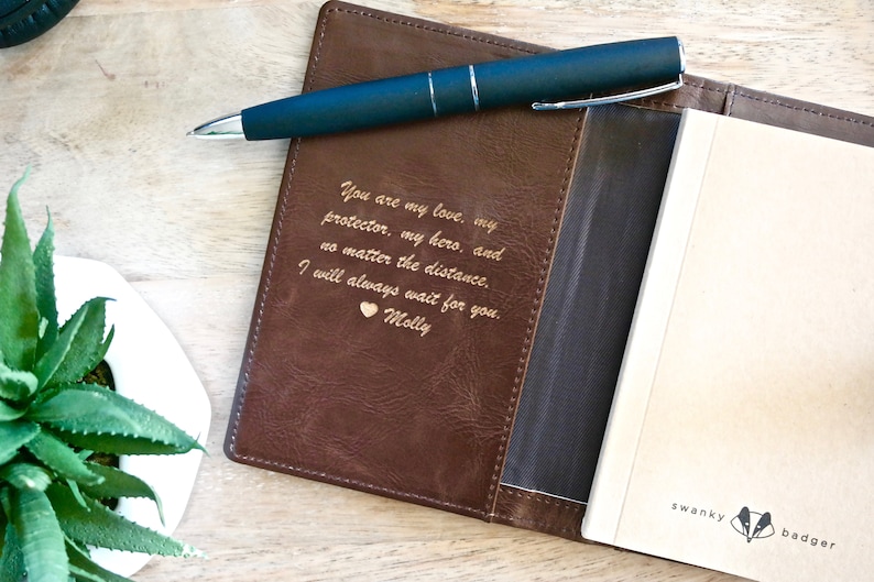 May include: A brown leather journal cover with a gold engraved message: "You are my love, my protector, my hero, and no matter the distance. I will always wait for you. Molly". The cover has a pocket for a pen and a black pen is placed in the pocket. The journal is open to a blank page with the words "swanky badger" printed on the bottom right corner.