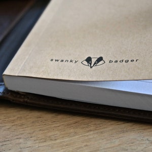 May include: A brown leather notebook cover with a white notebook inside. The cover has the text "swanky badger" and a badger silhouette.