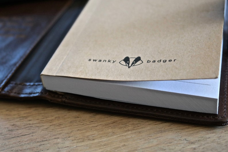 May include: A brown leather notebook cover with a white notebook inside. The cover has the text "swanky badger" and a badger silhouette.