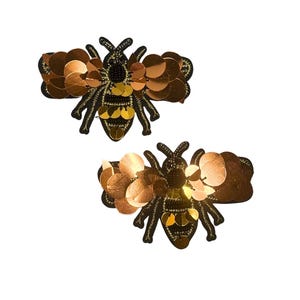 May include: Two bee-shaped patches featuring black embroidered bodies with gold and copper-colored circular sequins forming the wings and abdomen. The insect designs are detailed with black thread and metallic accents, creating a textured appearance.