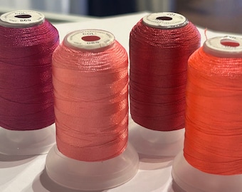 May include: Four spools of thread in shades of pink, red, and orange. Each spool is on a clear plastic base. The thread colors range from deep magenta to a vibrant orange-red. The spools are labeled with white text.