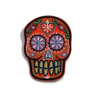 Puede incluir: Parche de calavera de azúcar bordada de color naranja con contornos negros, ojos morados y azules y un diseño floral.