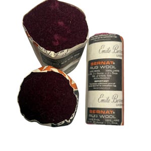 May include: Two bundles of deep burgundy rug wool yarn, each wrapped in a paper label. The labels feature the text Bernat Rug Wool, 100% Wool, 100% Laine, Net Wt. 2 1/2 Ounce, No. of Ends: 500, Moth-Proofed, and the name Emile Bernat.