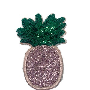 Può includere: Una toppa a forma di ananas rosa e verde con paillettes. L'ananas è rosa con glitter e le foglie sono paillettes verdi.