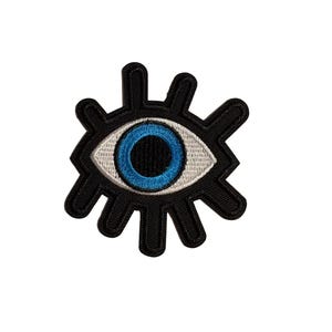 May include: An embroidered patch in the shape of an eye with a black outline and radiating black lashes. The eye features a white sclera, a bright blue iris, and a dark black pupil, creating a bold graphic design with a textured thread finish.
