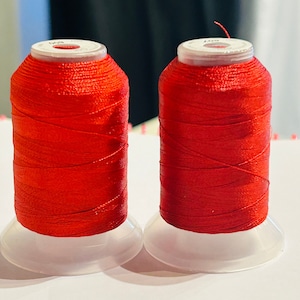 Puede incluir: Dos carretes de hilo rojo en bobinas de plástico blanco. El hilo está enrollado apretado y tiene un acabado brillante.