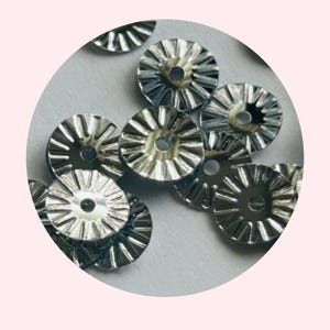 Puede incluir: Primer plano de lentejuelas redondas decorativas de color plateado con borde festoneado. Las lentejuelas tienen un agujero en el centro para coser o sujetar.