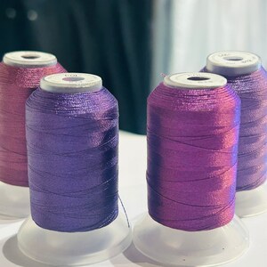 Puede incluir: Cuatro carretes de hilo en tonos de morado y rosa. Los carretes están dispuestos en fila sobre una superficie blanca.