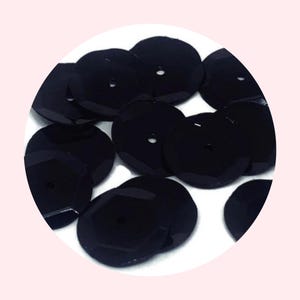 May include: A group of black round sequins with a hole in the center. The sequins are scattered on a white surface.