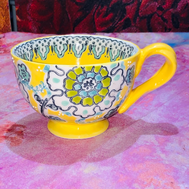 May include: A yellow ceramic teacup with a floral design. The cup has a large handle and a small pedestal base. The floral design includes blue, green, and white accents on a yellow background. The rim of the cup has a decorative pattern.