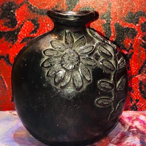 May include: A black, round vase with a detailed floral design carved into its surface. The flower and leaf pattern is raised, creating a textured effect. The vase has a small, rounded neck and a dark, glossy finish.