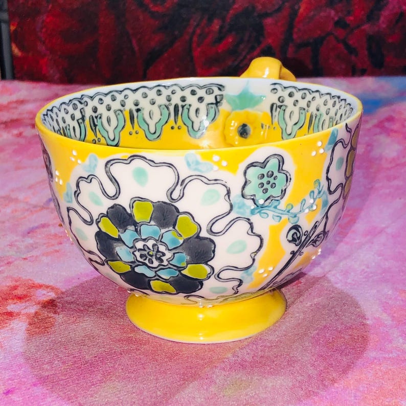 May include: A yellow ceramic bowl with a floral design. The bowl has a pedestal base and a handle. The exterior is decorated with large flowers in shades of blue, green, and grey against a yellow background.