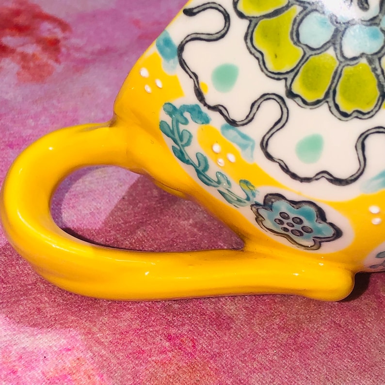 May include: A yellow ceramic teacup with a curved handle. The cup has a floral design in white, green, and blue. The handle is bright yellow, and the cup is set against a pink background.