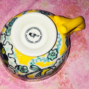 May include: A yellow ceramic teacup with a floral design in blue, black, and white. The handle is a bright yellow, and the bottom of the cup has a white interior with a bird logo and text that reads "Microwave & Dishwasher Safe Made in China".