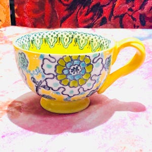 May include: A yellow teacup with a matching handle and base. The cup features a floral design in blue, green, and yellow on a white background. The rim has a decorative green and white pattern. The interior is bright yellow.