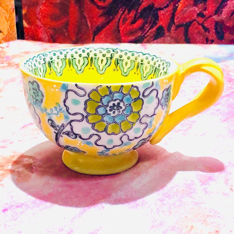 May include: A yellow teacup with a matching handle and base. The cup features a floral design in blue, green, and yellow on a white background. The rim has a decorative green and white pattern. The interior is bright yellow.