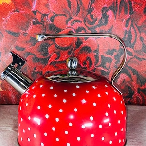 May include: A red tea kettle with white polka dots. The kettle has a silver spout, lid, and handle. The handle arches over the top of the kettle. The background is a red floral pattern.