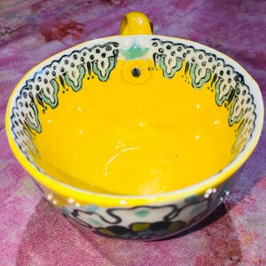 May include: A bright yellow ceramic bowl with a small handle. The rim has a decorative pattern of black, white, and green floral designs. The interior is a solid, glossy yellow. A small yellow flower is painted on the inside of the bowl.