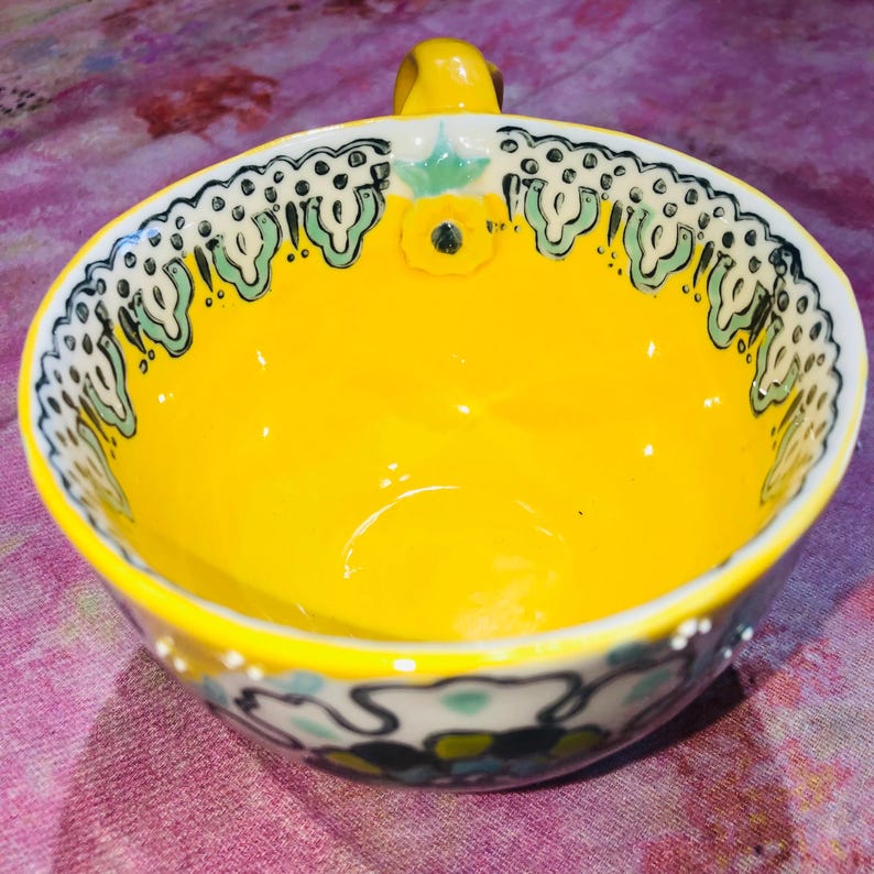 May include: A bright yellow ceramic bowl with a small handle. The rim has a decorative pattern of black, white, and green floral designs. The interior is a solid, glossy yellow. A small yellow flower is painted on the inside of the bowl.