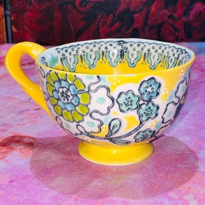 May include: A yellow teacup with a matching handle and base. The cup is decorated with a floral pattern in shades of blue, green, and white against a yellow background. The rim features a scalloped design.