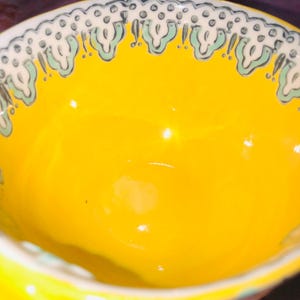 May include: A vibrant yellow ceramic bowl with a patterned rim. The rim has a repeating design of white, pale green, and grey shapes. The bowl's interior is a solid, glossy yellow.