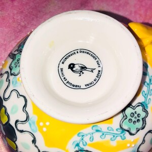 May include: A white ceramic bowl with a yellow and floral design. The base of the bowl features a black circular stamp with a bird illustration and the text "MICROWAVE & DISHWASHER SAFE MADE IN CHINA FABRIQUE EN CHINE".