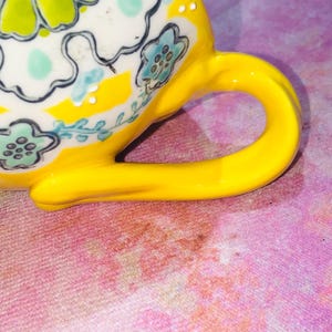 May include: Close-up of a yellow ceramic teacup with a curved handle. The cup features a floral design with blue and white accents on a yellow background. The handle is a bright, solid yellow.