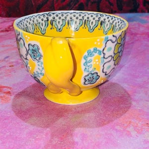 May include: A yellow ceramic teacup with a bird-shaped handle and a small foot. The cup features a floral design in blue, white, and yellow. The rim has a decorative pattern. The cup is approximately 10 cm in diameter.