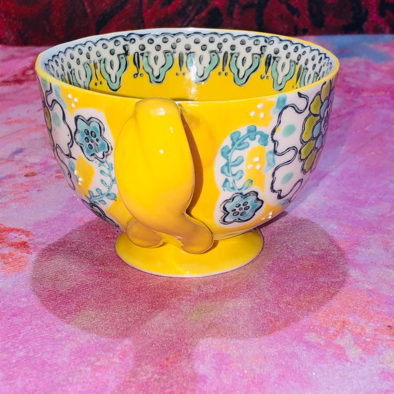 May include: A yellow ceramic teacup with a bird-shaped handle and a small foot. The cup features a floral design in blue, white, and yellow. The rim has a decorative pattern. The cup is approximately 10 cm in diameter.