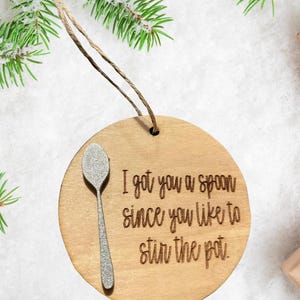 May include: Wooden Christmas ornament with a silver spoon and the engraved text: "I got you a spoon since you like to stir the pot." The ornament is round and has a jute string for hanging.