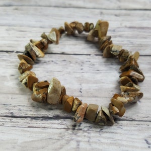 May include: A bracelet made of irregularly shaped, light brown and beige jasper stones. The stones are strung together to form a flexible band. The bracelet is displayed on a light-colored, wooden surface.
