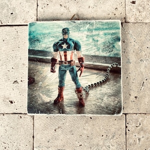 Könnte beinhalten: Ein quadratisches, verwittertes Kunstwerk mit einer Captain America-Actionfigur. Die Figur trägt ein blaues, weißes und rotes Kostüm mit einem Stern auf der Brust. Der Hintergrund hat ein strukturiertes, verwittertes Aussehen.