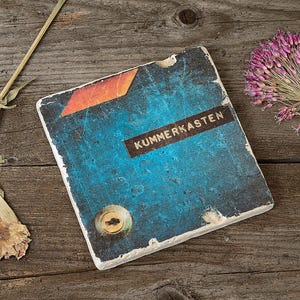 Könnte beinhalten: Ein verwittertes, quadratisches, blau-oranges Buch mit dem Wort "KUMMERKASTEN" auf einem dunklen Etikett. Eine getrocknete Blume und eine lila Alliumblüte liegen auf einer Holzoberfläche.