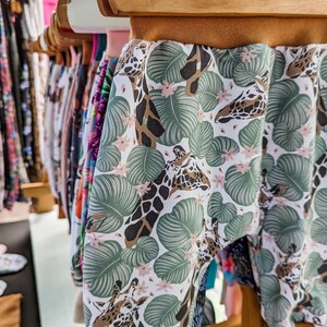 May include: Pants featuring a white background with a pattern of green tropical leaves, small pink flowers, and brown giraffe heads. The garment has a solid brown elastic waistband and matching brown elastic cuffs at the bottom of the legs.